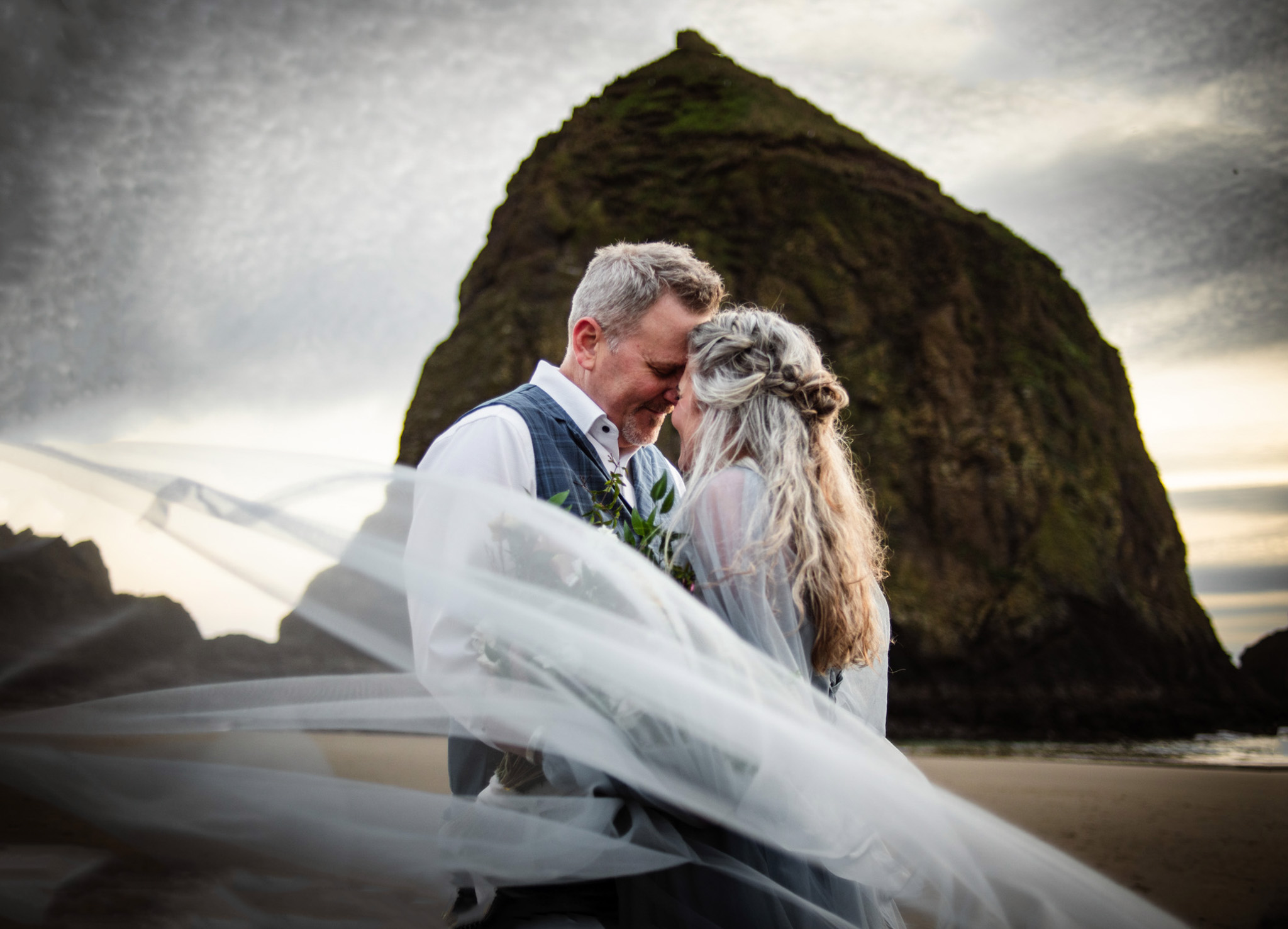 Couple in wedding attire standing on beach with large rock formation in background, wedding veil flowing, overcast sky