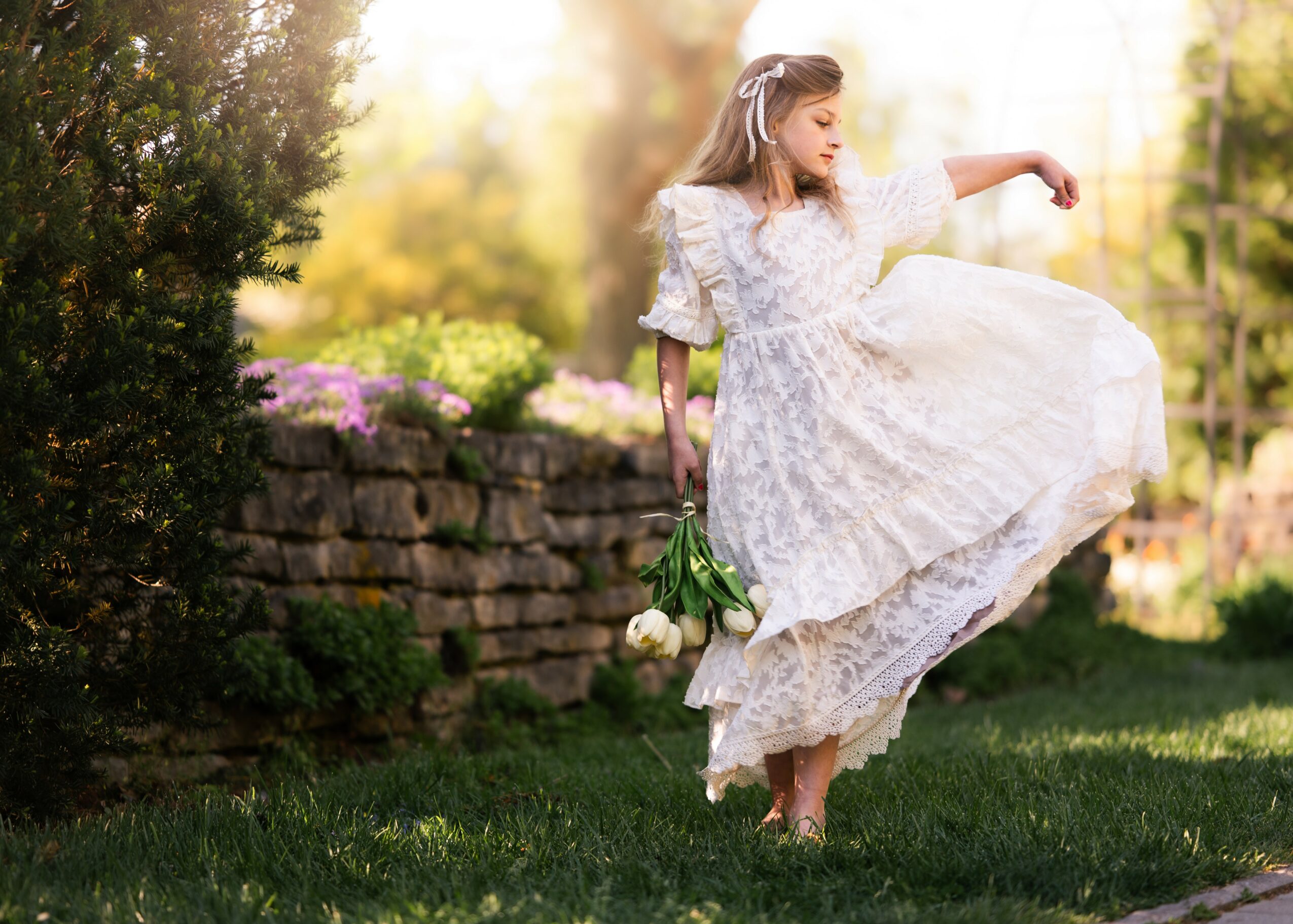 Girl in a white dress holding flowers, lifting her dress while walking outdoors in sunlight.