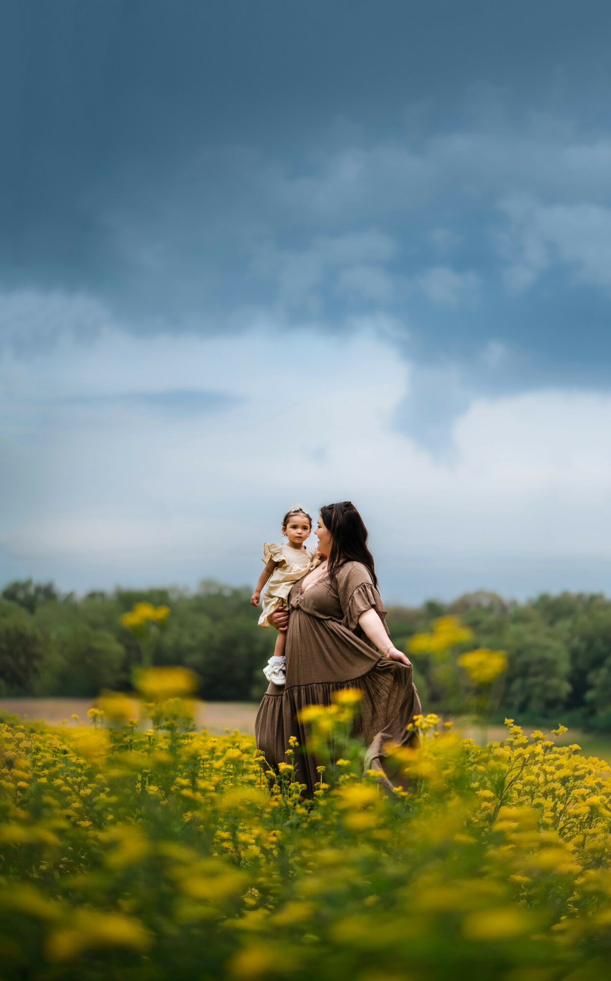 A woman carrying a child in a field of yellow flowers under a cloudy sky.
