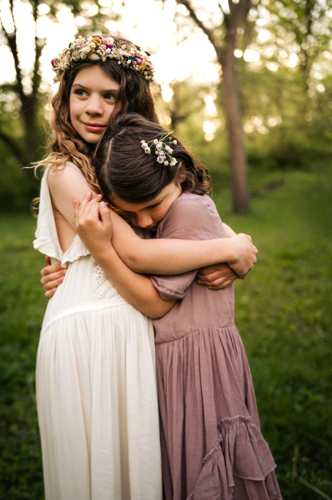 Two young girls hugging outdoors, one wearing a white dress and floral crown, the other in a mauve dress, in a green park.