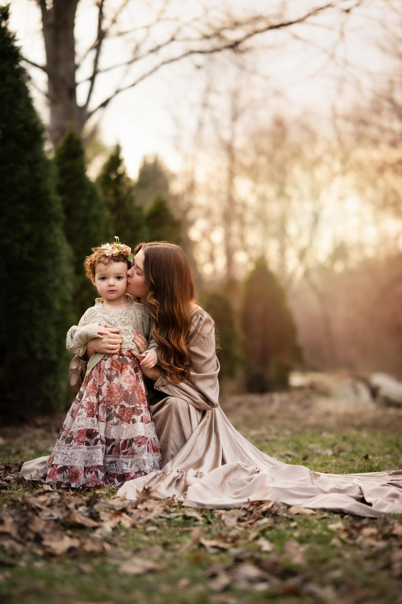 Woman kneeling with a young girl outdoors, trees and sunlight in background, autumn setting, girl in floral dress, woman in long dress.