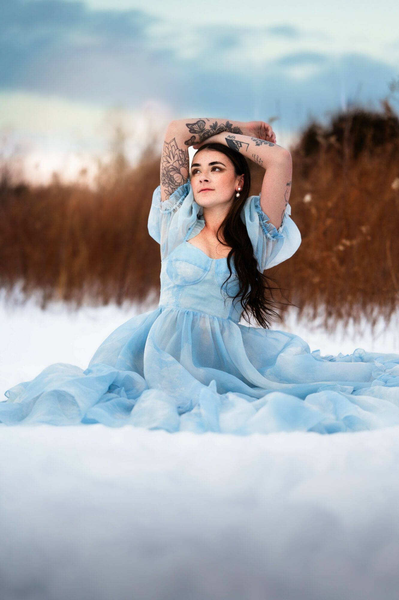 Woman in a light blue dress sitting on snow with arms raised above her head outdoors, sky and trees in background.