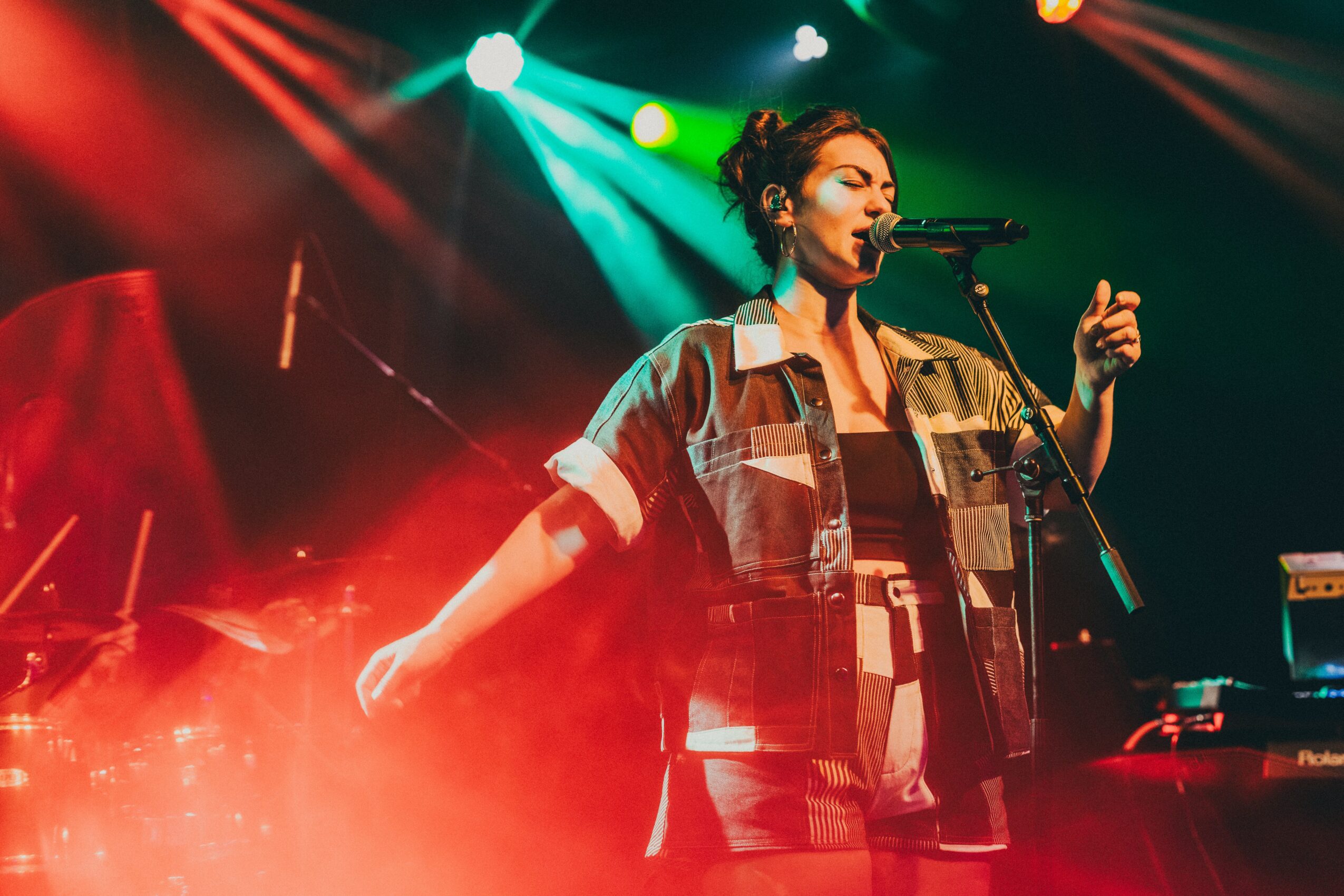 Female singer performing on stage with colorful lights, wearing a plaid shirt and gesturing with her hand.