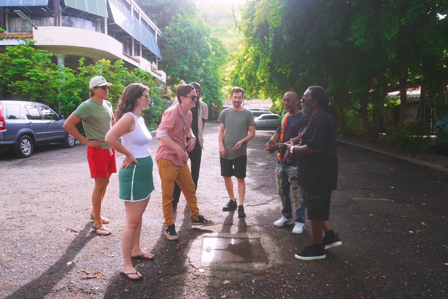 Group of seven people standing outdoors in a circle, talking and smiling, with trees and parked cars in the background.