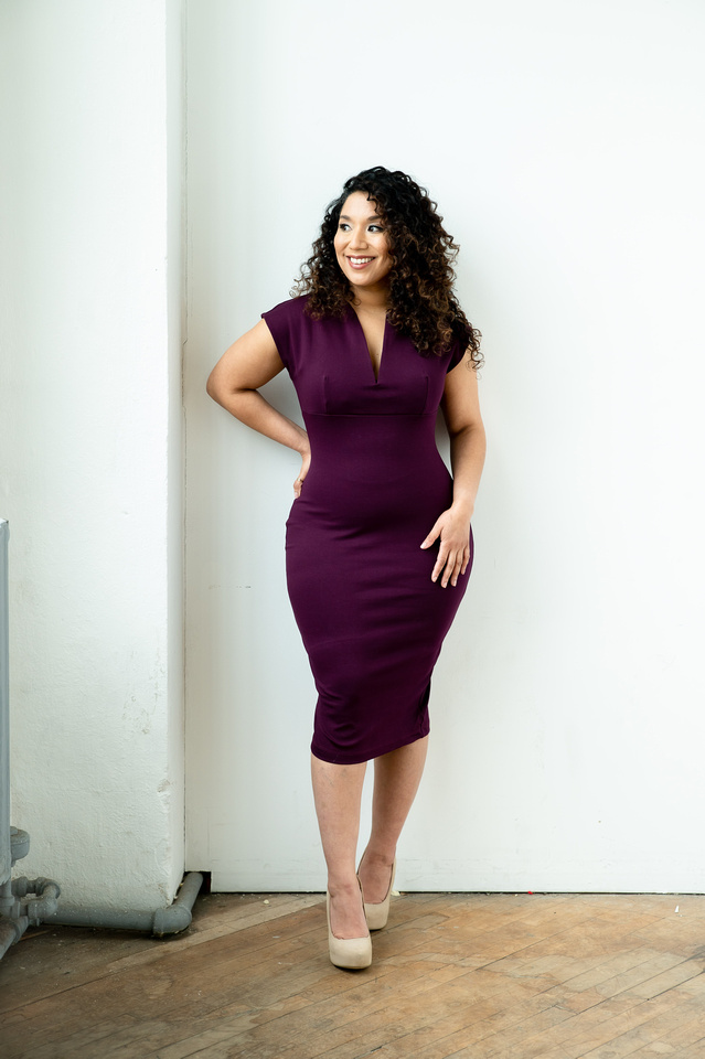 Woman with curly hair in a purple dress standing indoors near a white wall, smiling, with hands on hips, wearing beige heels.
