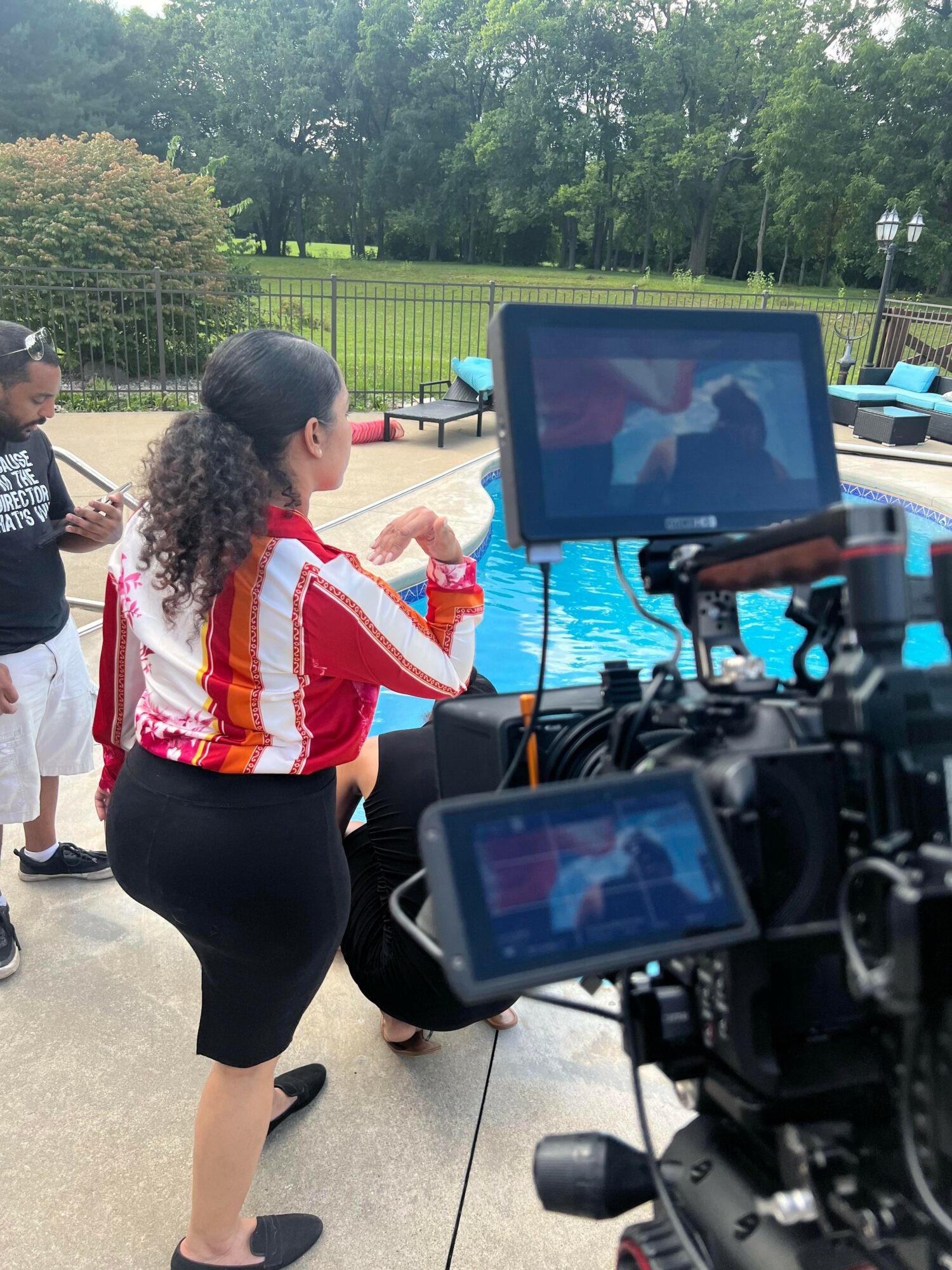 People gathered outdoors near a pool with a camera recording a woman in a red and white striped shirt, black shorts, and curly hair.