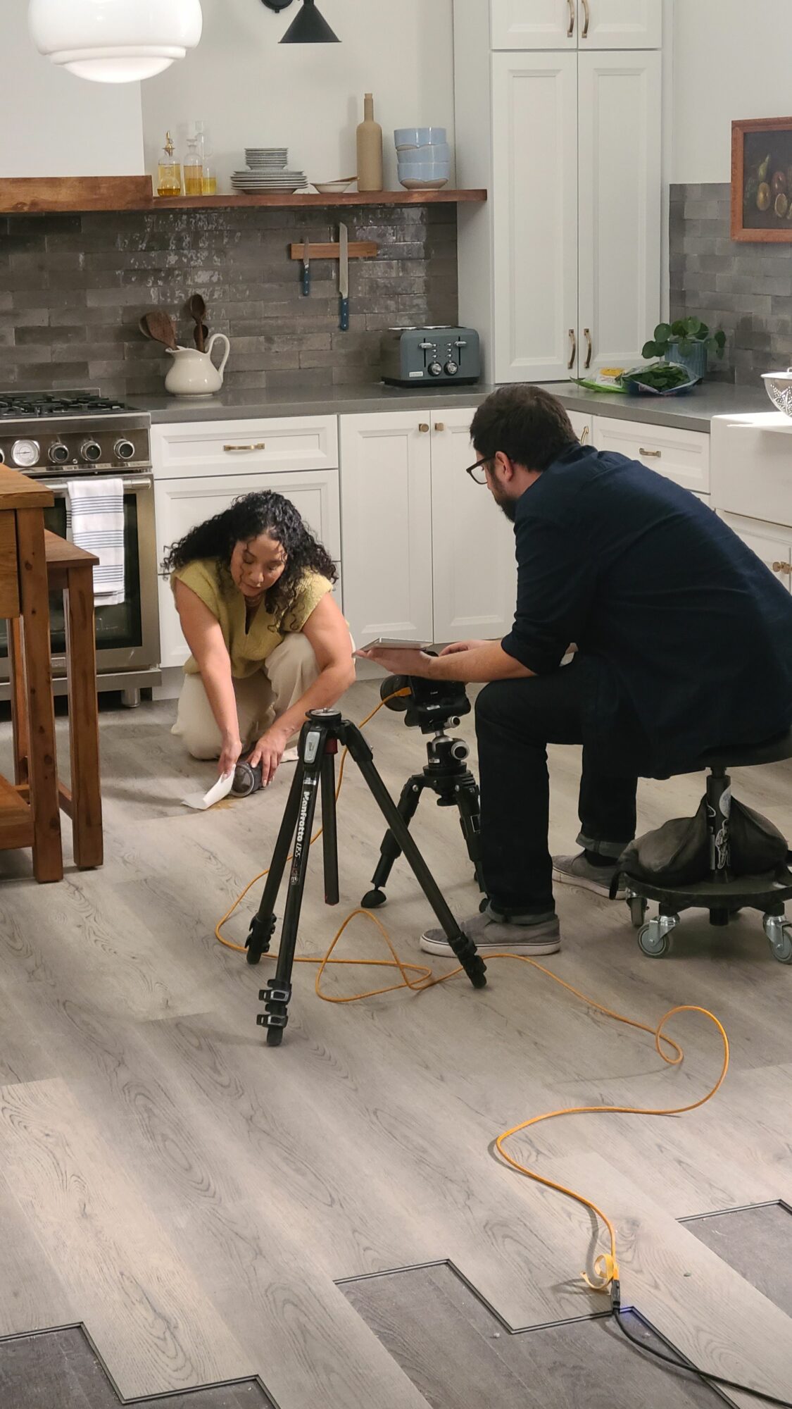 Person adjusting camera equipment in a room with a woman sitting on the floor, kitchen in background.