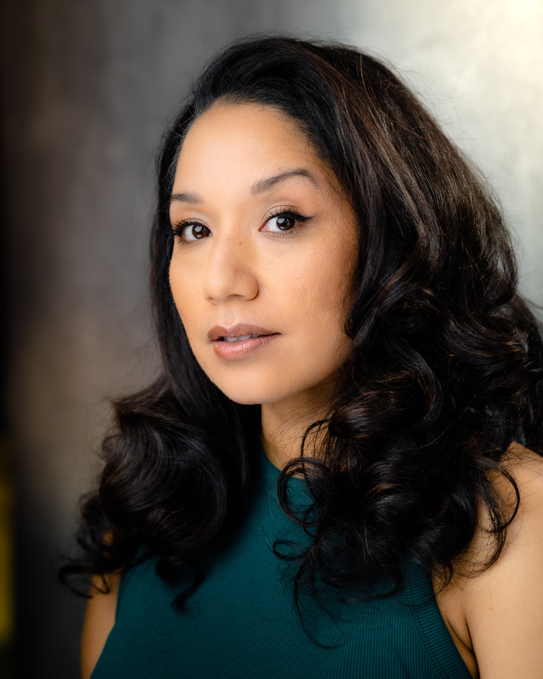 Portrait of a woman with long, dark, wavy hair wearing a teal top, looking at the camera with a neutral expression.