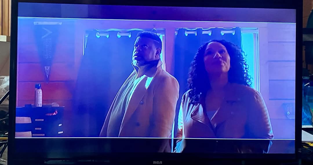 Two people stand side by side in front of blue-lit curtains, facing forward, in a room with wooden walls.