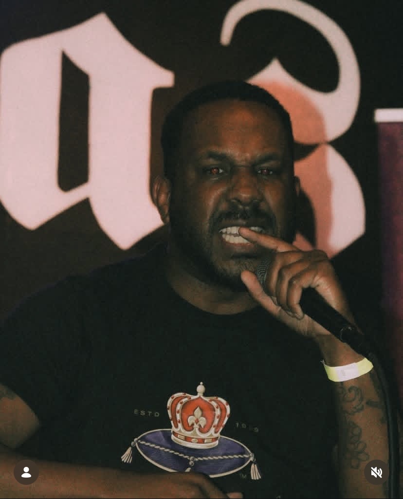Person with dark hair and beard holding a microphone, wearing a black shirt, in front of a dark background with white lettering.