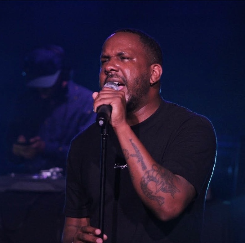 Man singing into a microphone with a serious expression, dark background, and a person in the background wearing a cap.