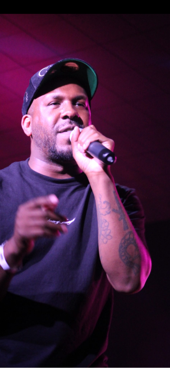 Man holding microphone, wearing a cap, performing on stage with pink lighting, focused expression, dark background.