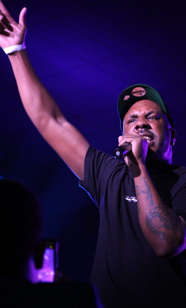 Male performer singing into microphone with one arm raised, wearing a cap and black shirt, stage lighting in background.