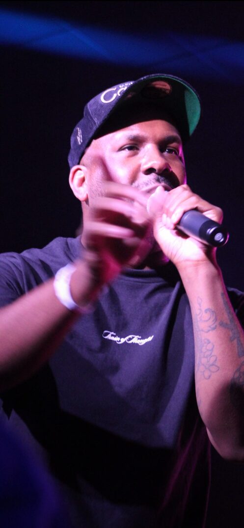 Person wearing a cap and black shirt, holding a microphone, speaking or performing on stage, with dark background.