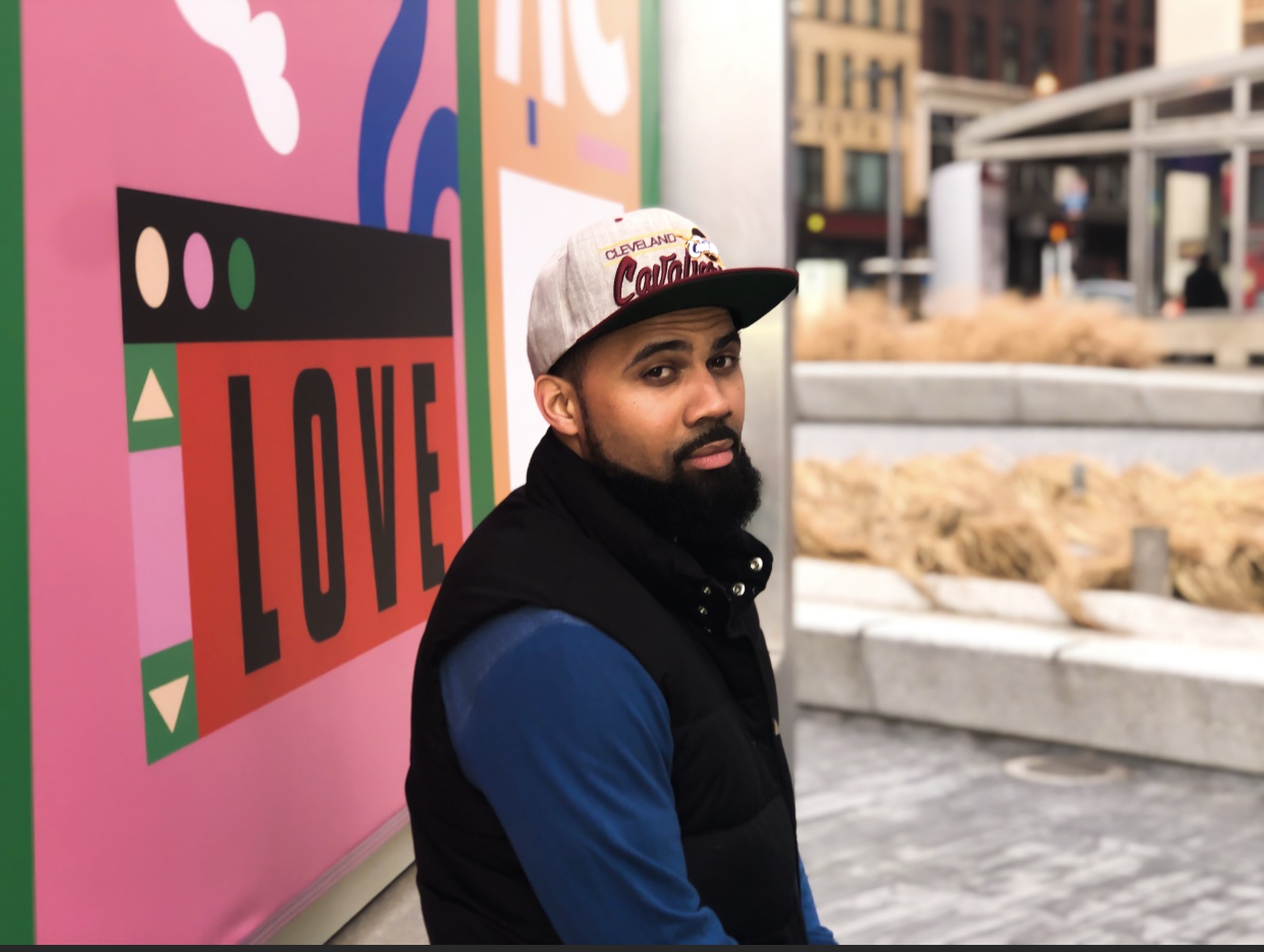 Young man with a beard wearing a gray cap and black vest, sitting outdoors near a colorful wall with the word 'LOVE'.