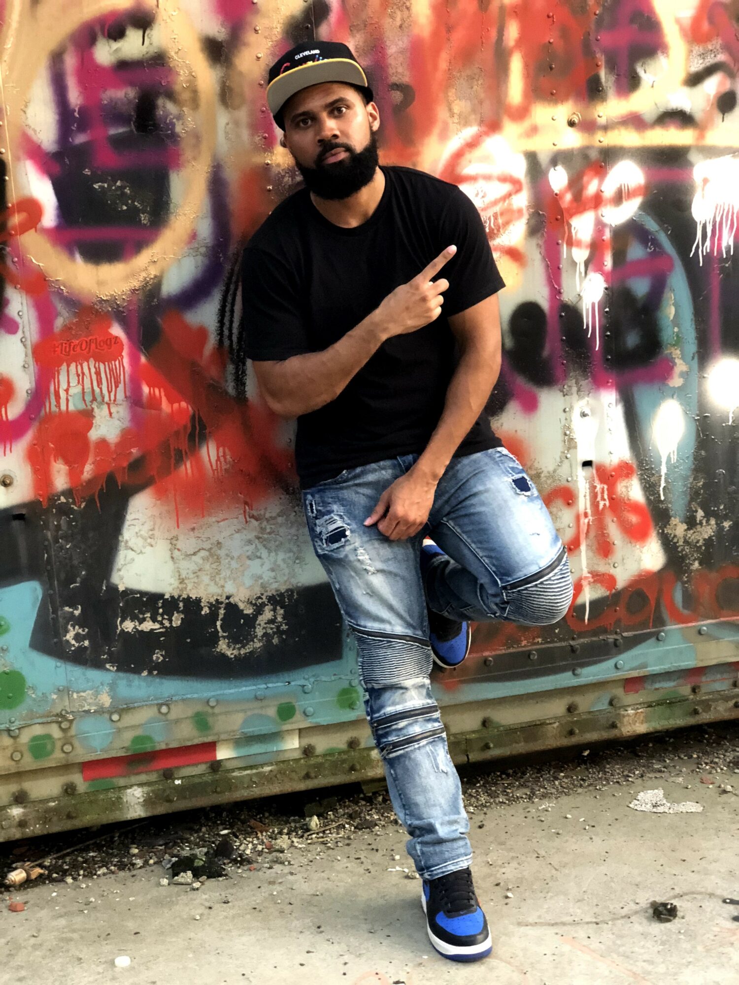 Man with beard and hat leaning against graffiti-covered wall, pointing upward with one finger, wearing black t-shirt and ripped jeans.
