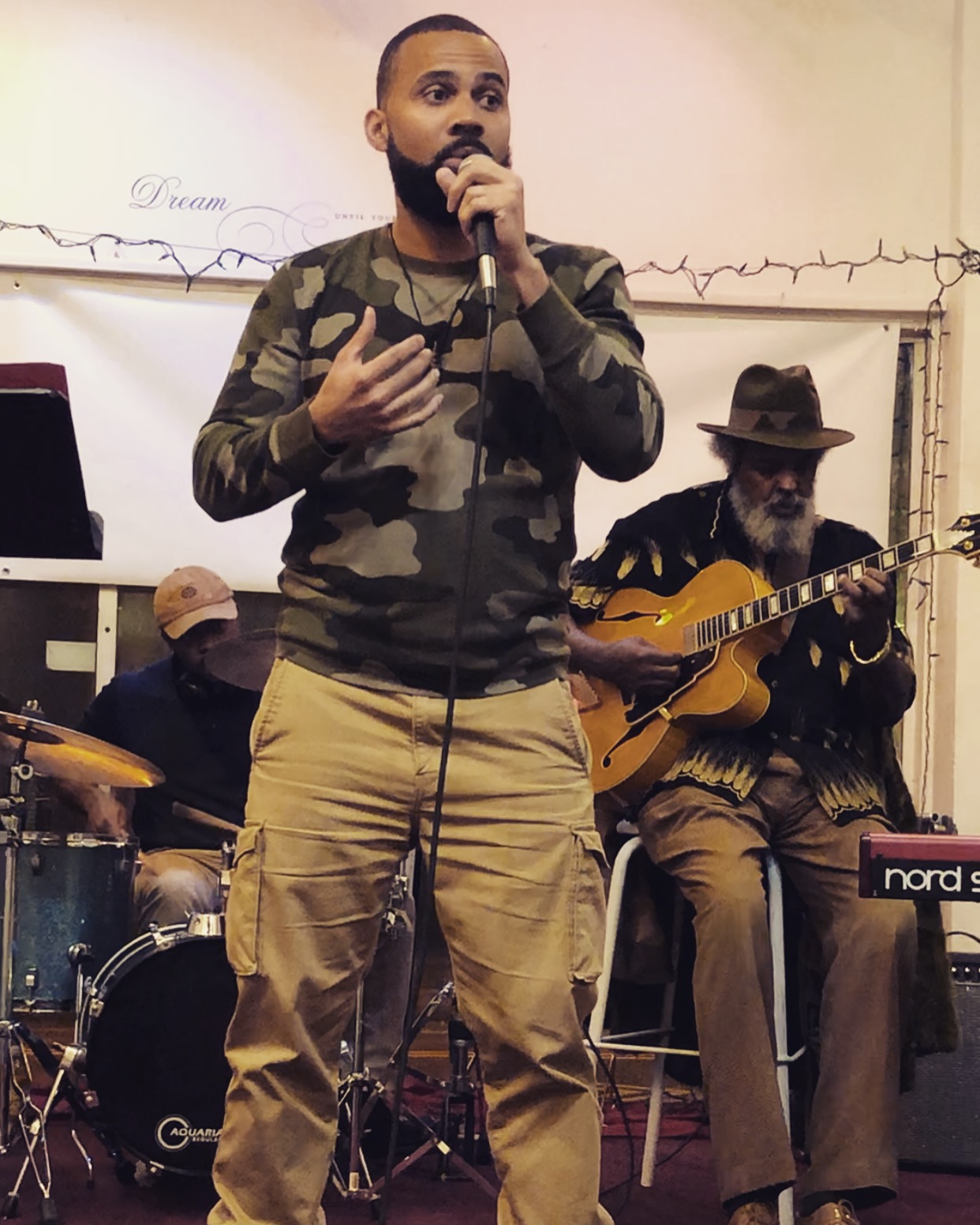 Man singing into microphone with band members playing instruments behind him, in an indoor setting.