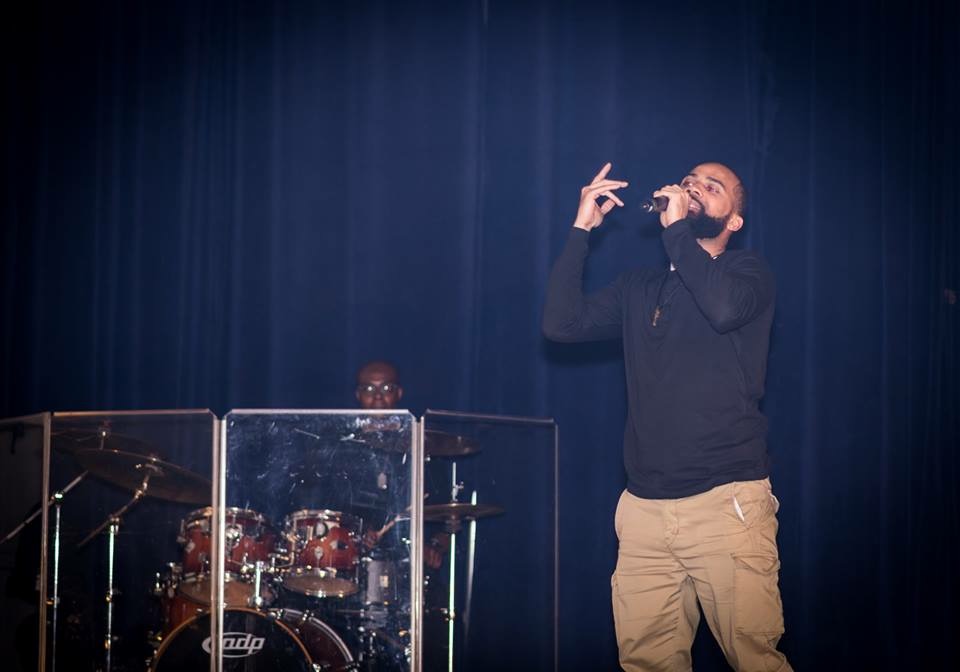 A man singing into a microphone on stage with a drummer behind a transparent shield, dark blue curtain backdrop.
