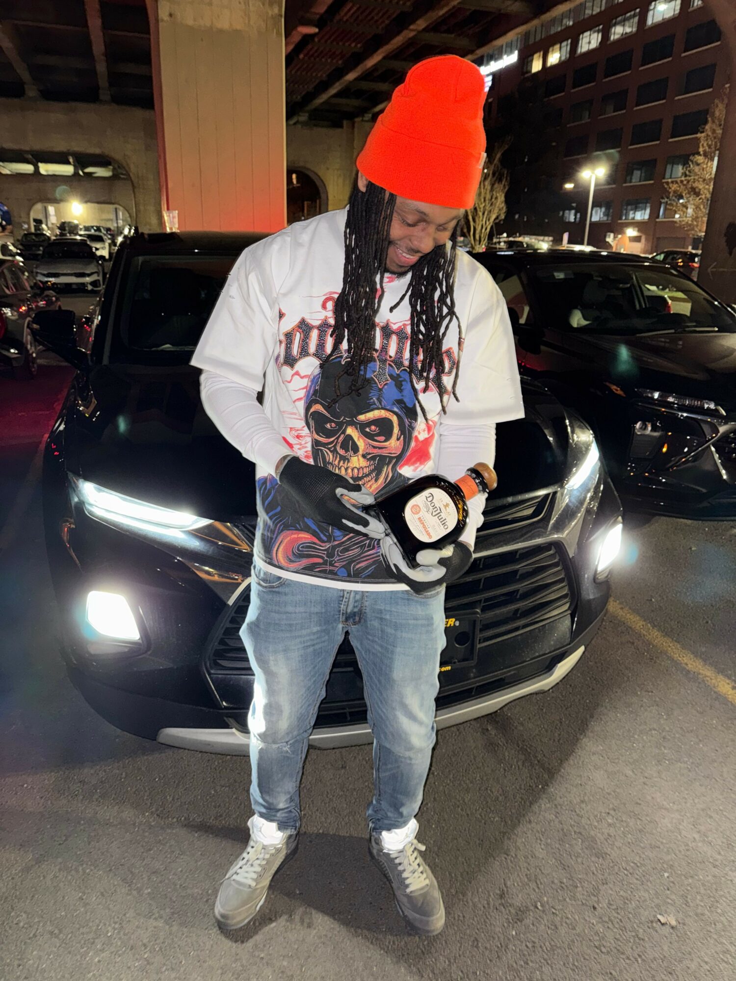 Person with long braided hair, wearing a white jacket, light blue jeans, and gray shoes, standing in a parking lot at night.