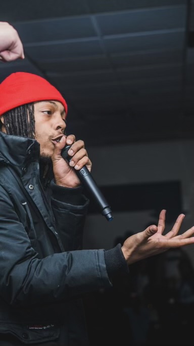 Person wearing a red hat and black jacket holding a microphone, gesturing with their hand, indoors.