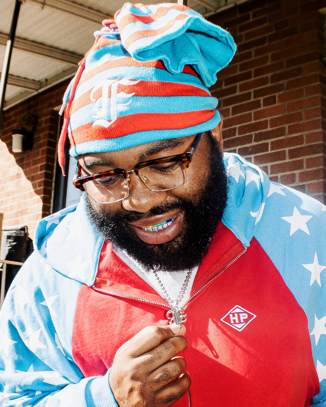 Man with glasses and a beard wearing a colorful hoodie and a striped hat, looking down, outdoors with brick wall background.