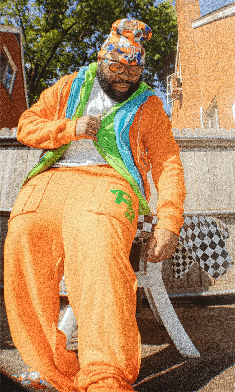 Person wearing colorful clothing, including orange pants and a multicolored hat, outdoors near a table with checkered tablecloth.