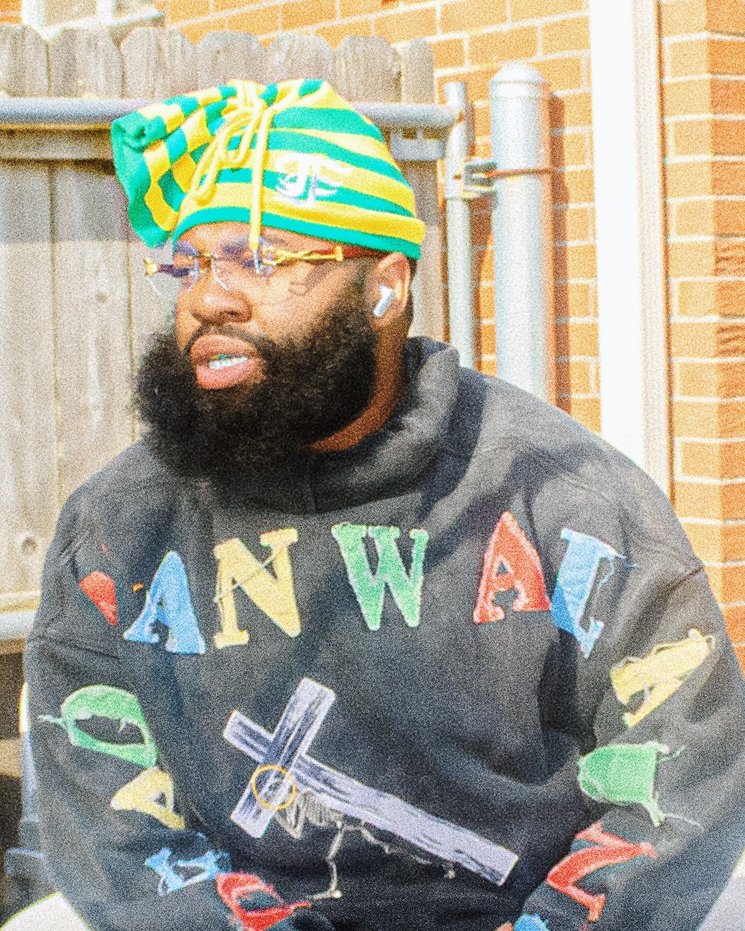 Man with a beard wearing glasses, a colorful striped headscarf, and a dark hoodie with colorful letters and a sword illustration, standing outdoors.