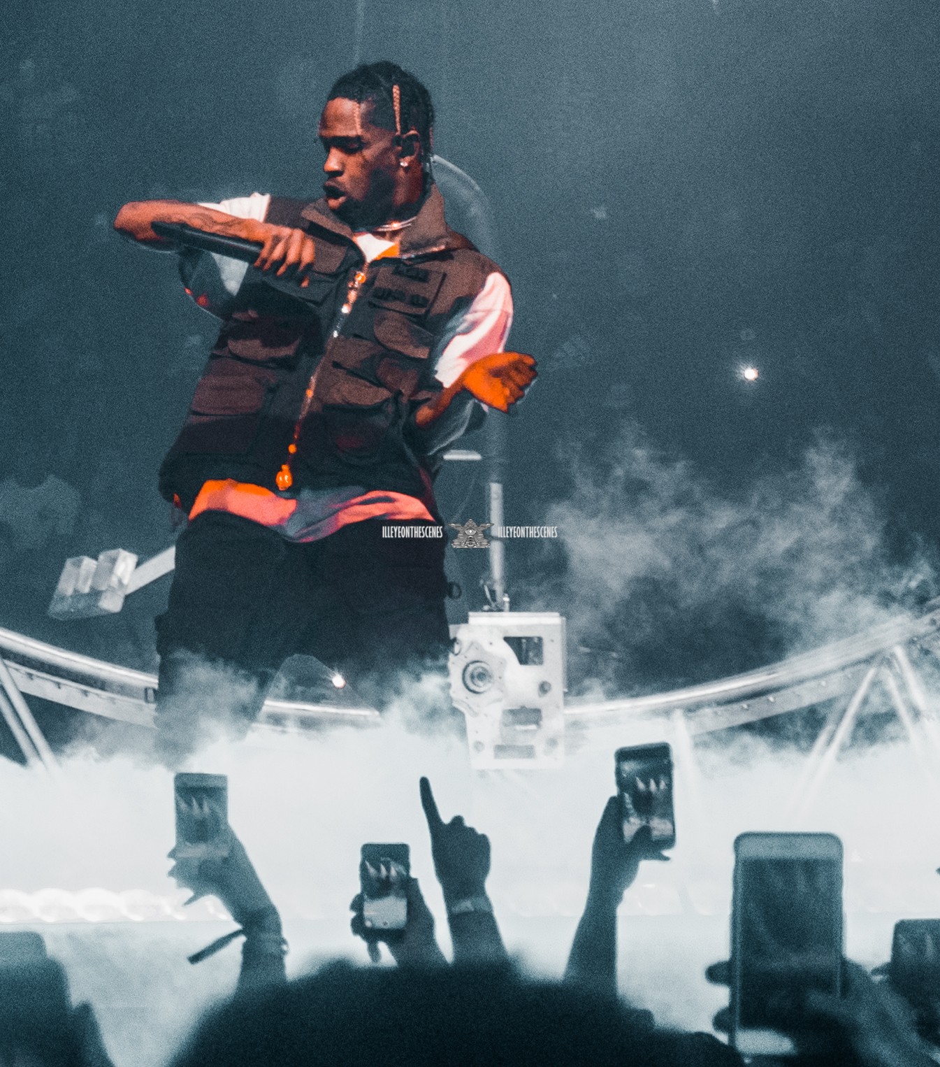 Performer on stage with smoke effects, holding a microphone, audience members capturing with phones, dark background.