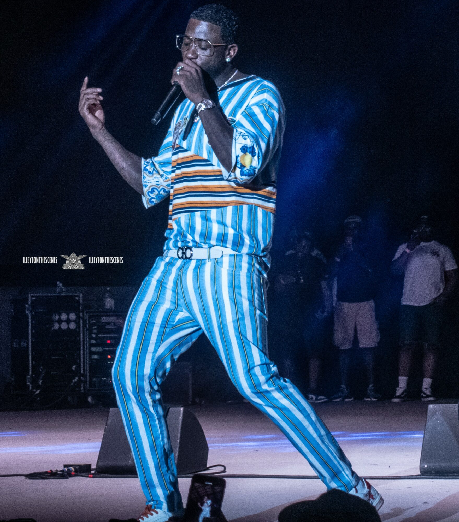 Performer singing into microphone on stage, wearing blue and white striped outfit, with people in background.