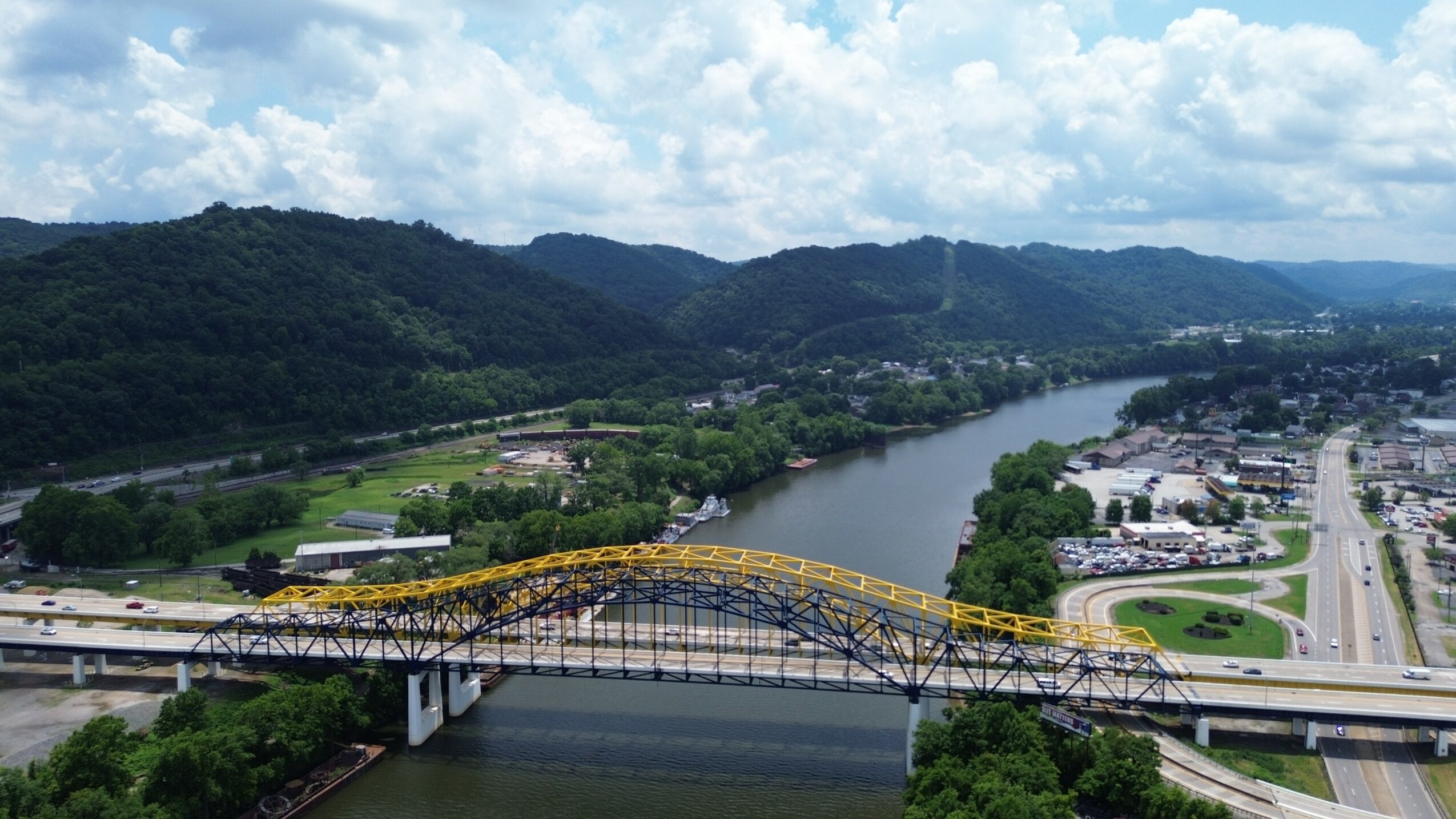A yellow bridge spans a river with a city and green hills in the background.