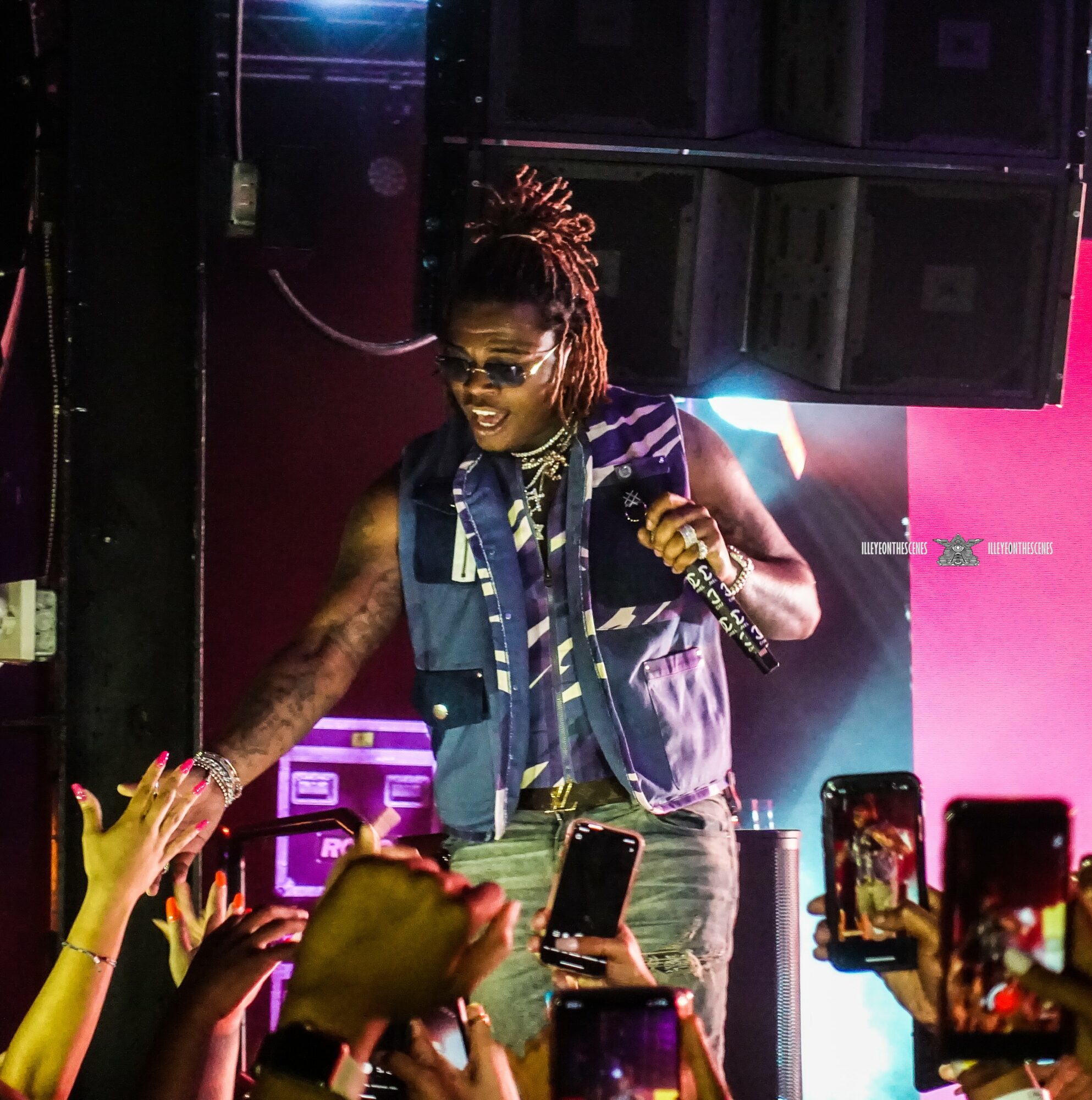 Performer with dreadlocks, sunglasses, and denim vest on stage, engaging with audience holding phones, colorful lighting in background.