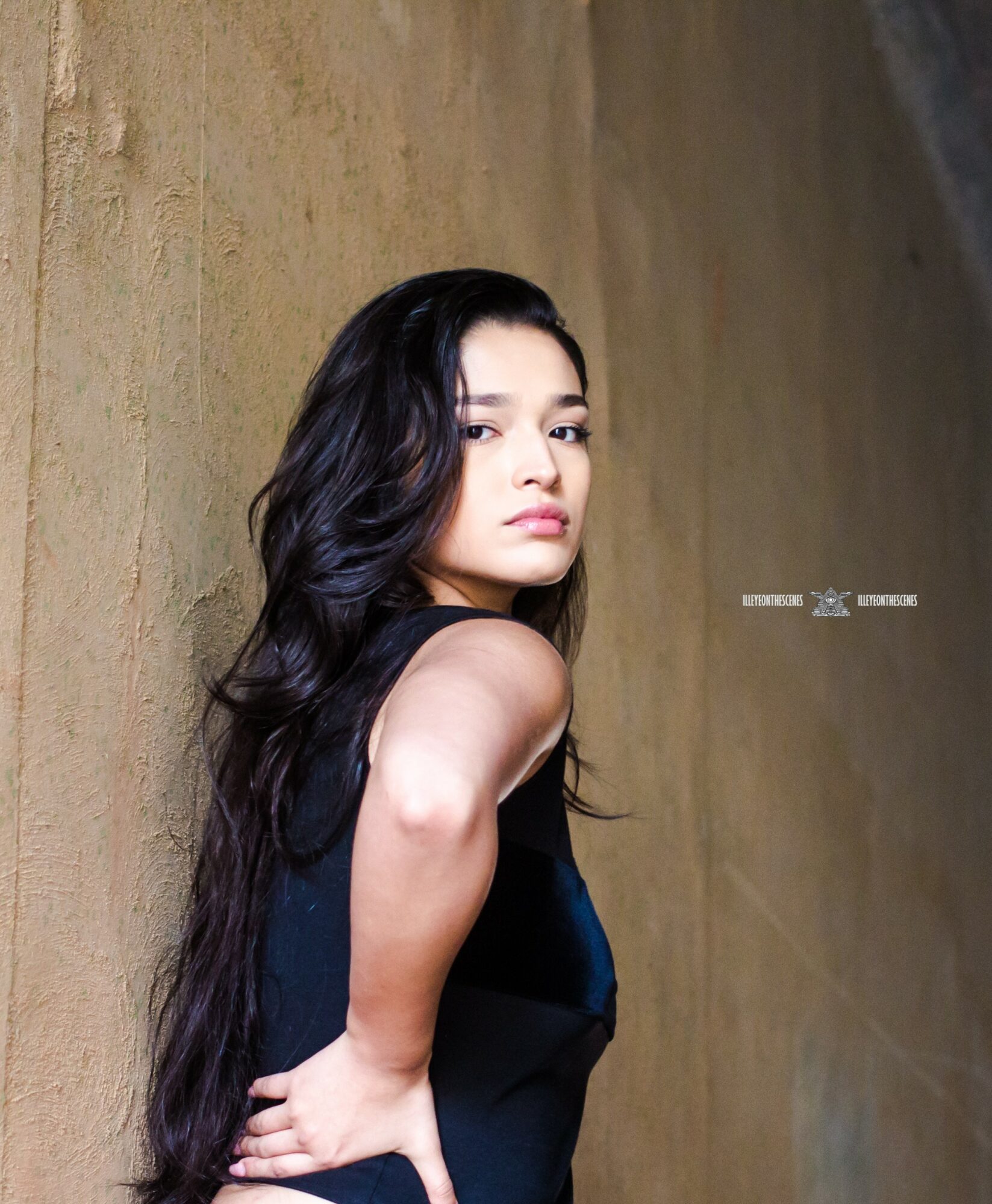 Young woman with long dark hair poses against a textured wall, looking at the camera with a neutral expression.
