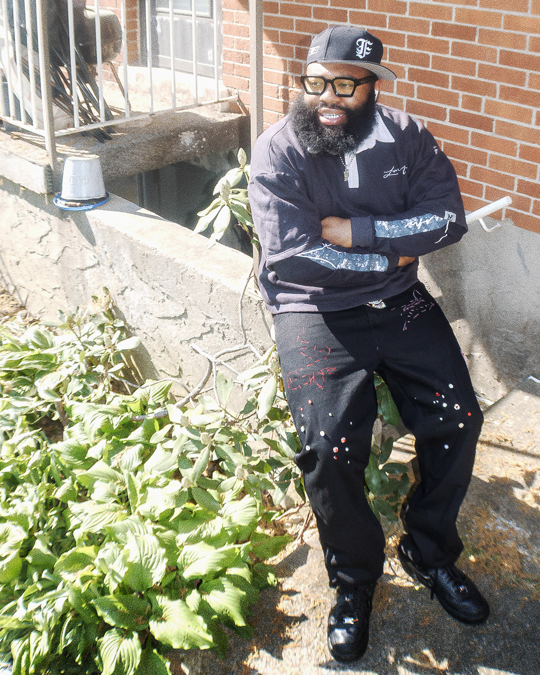 Man with glasses and a beard, wearing a cap and dark clothing, standing outdoors near a brick wall and plants.