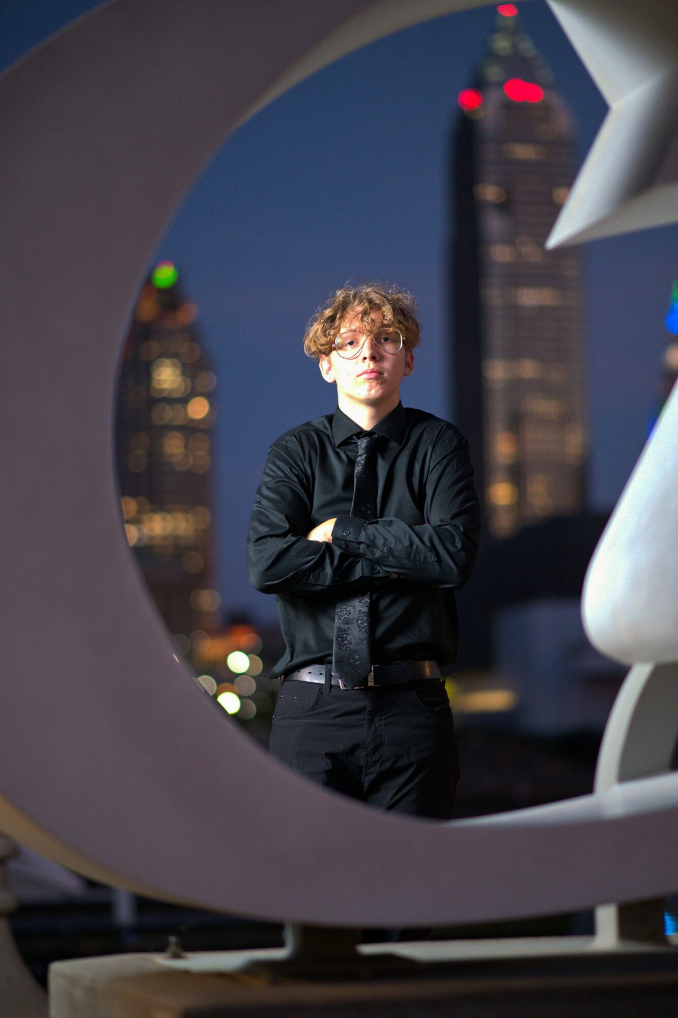 Young man with curly hair and glasses stands with arms crossed against a cityscape at night, framed by modern architectural elements.