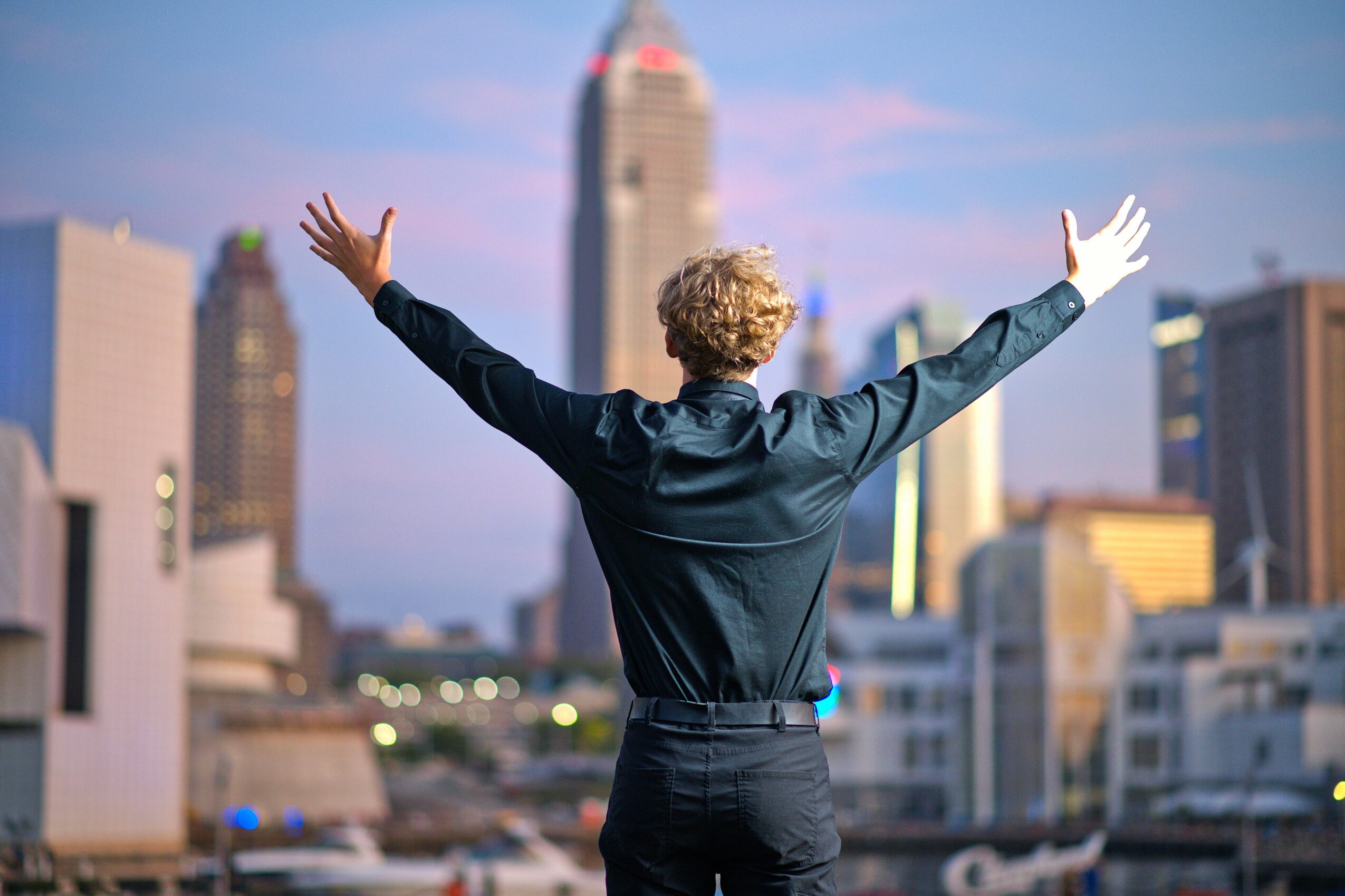 Person with curly hair and dark clothing stands with arms raised in front of a city skyline.