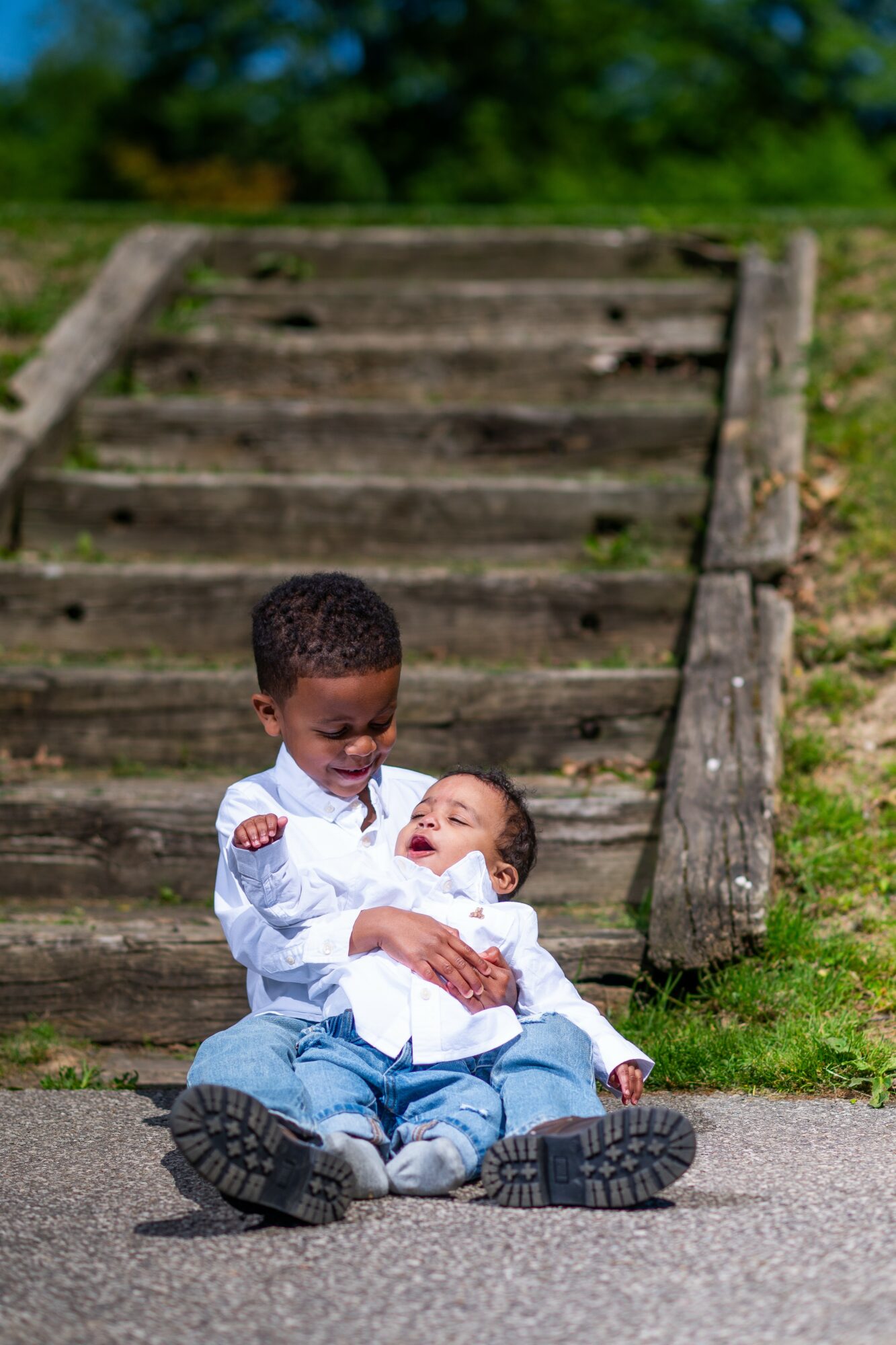Two children sitting on the ground in front of wooden stairs outdoors, one child holding the other, smiling.