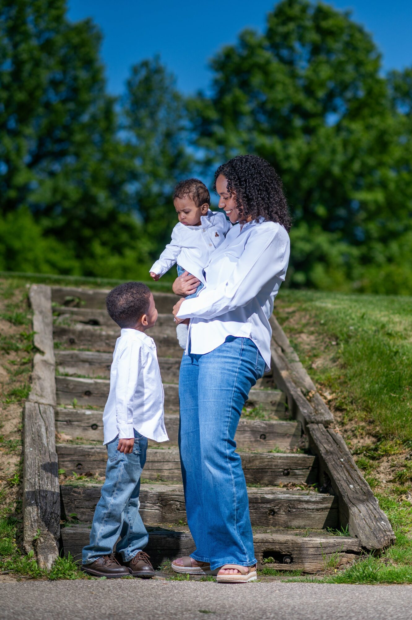 Woman holding a child while a boy stands on stairs outdoors with trees and blue sky in background.