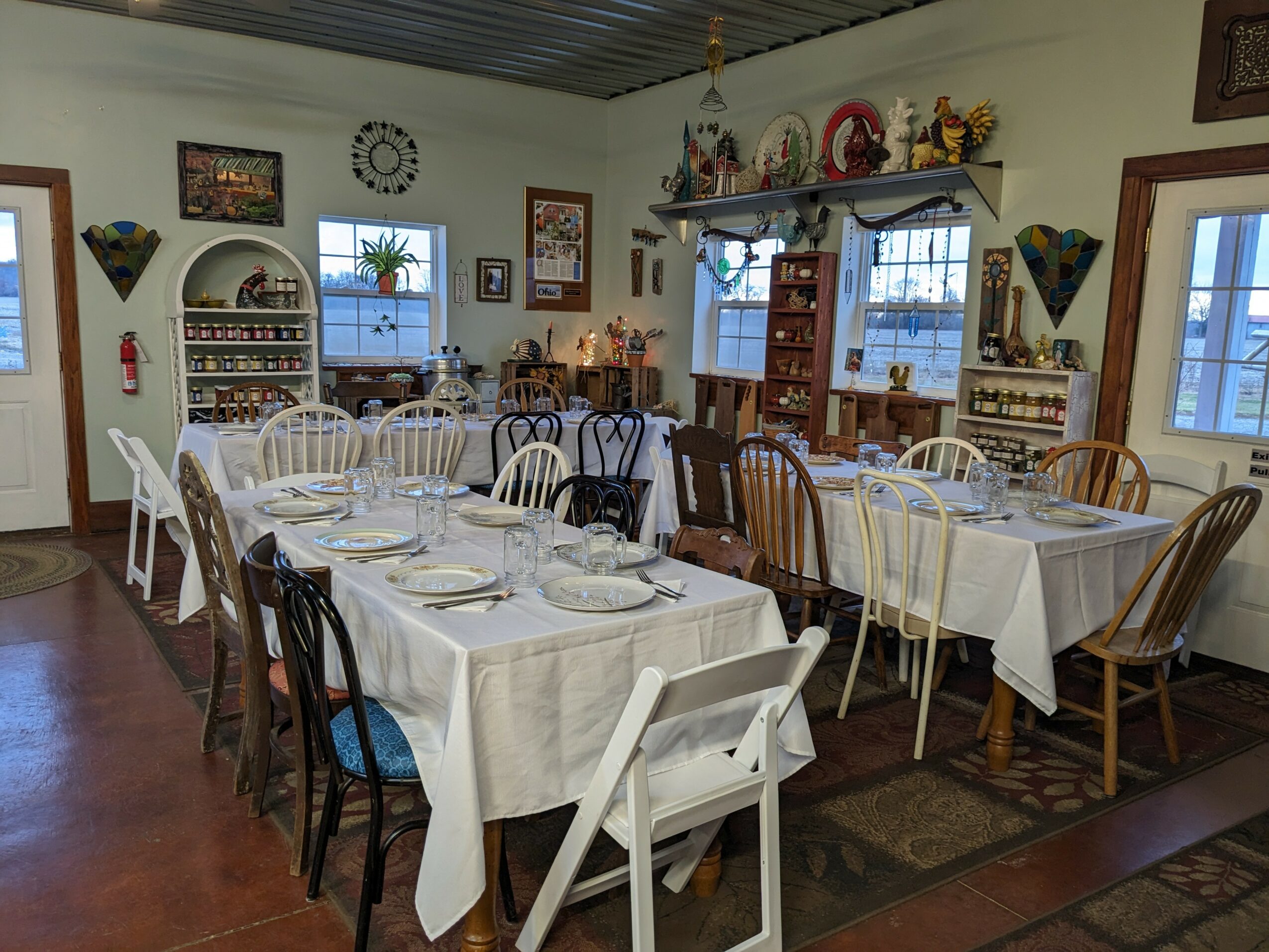 Dining room with tables set for a meal, white and wooden chairs, shelves with decorative items, and windows with curtains.