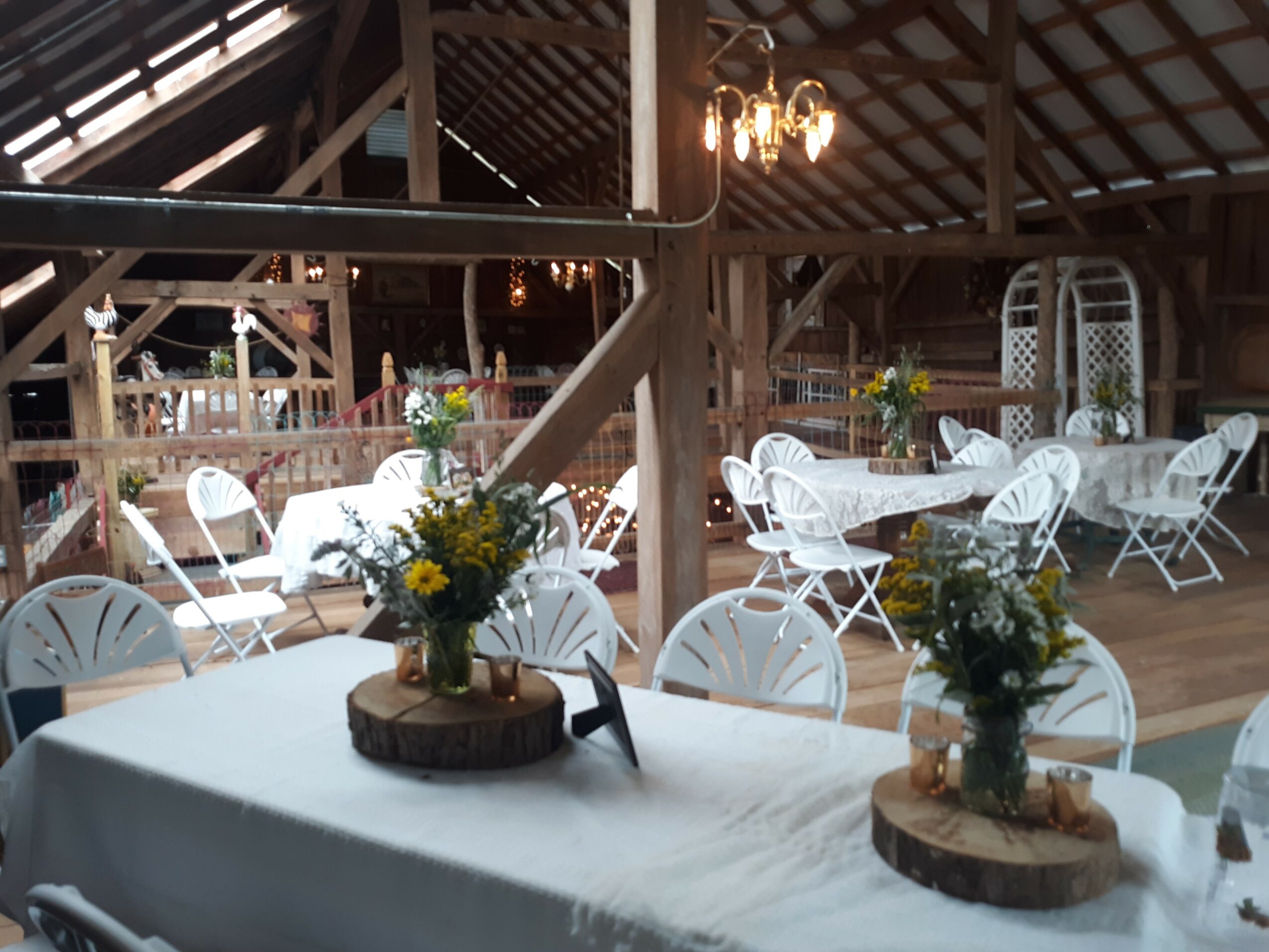 Event space with round tables, white chairs, floral centerpieces, and wooden accents in a barn-like setting.