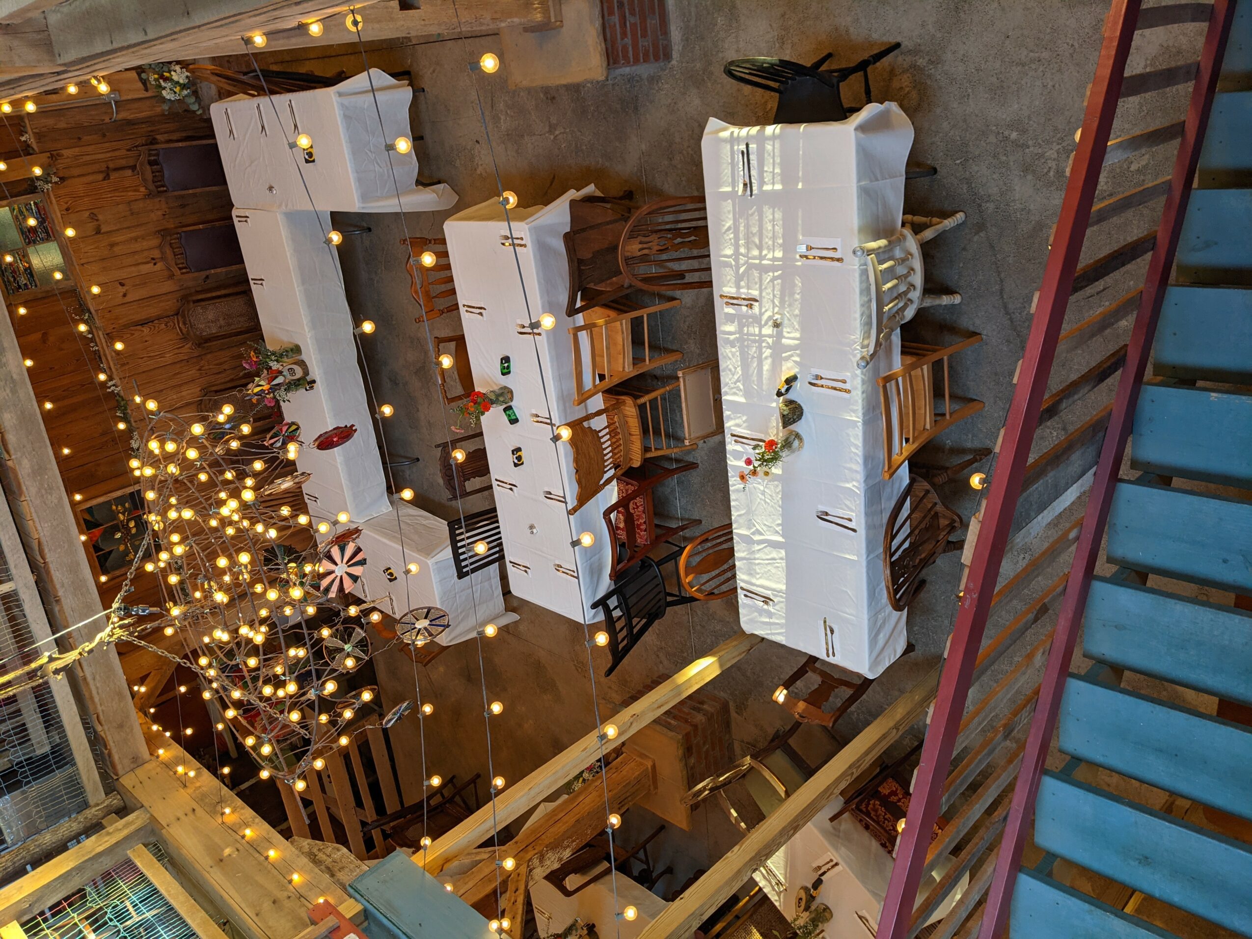 Indoor event space with decorated tables, string lights, and a tree with lights, viewed from above.