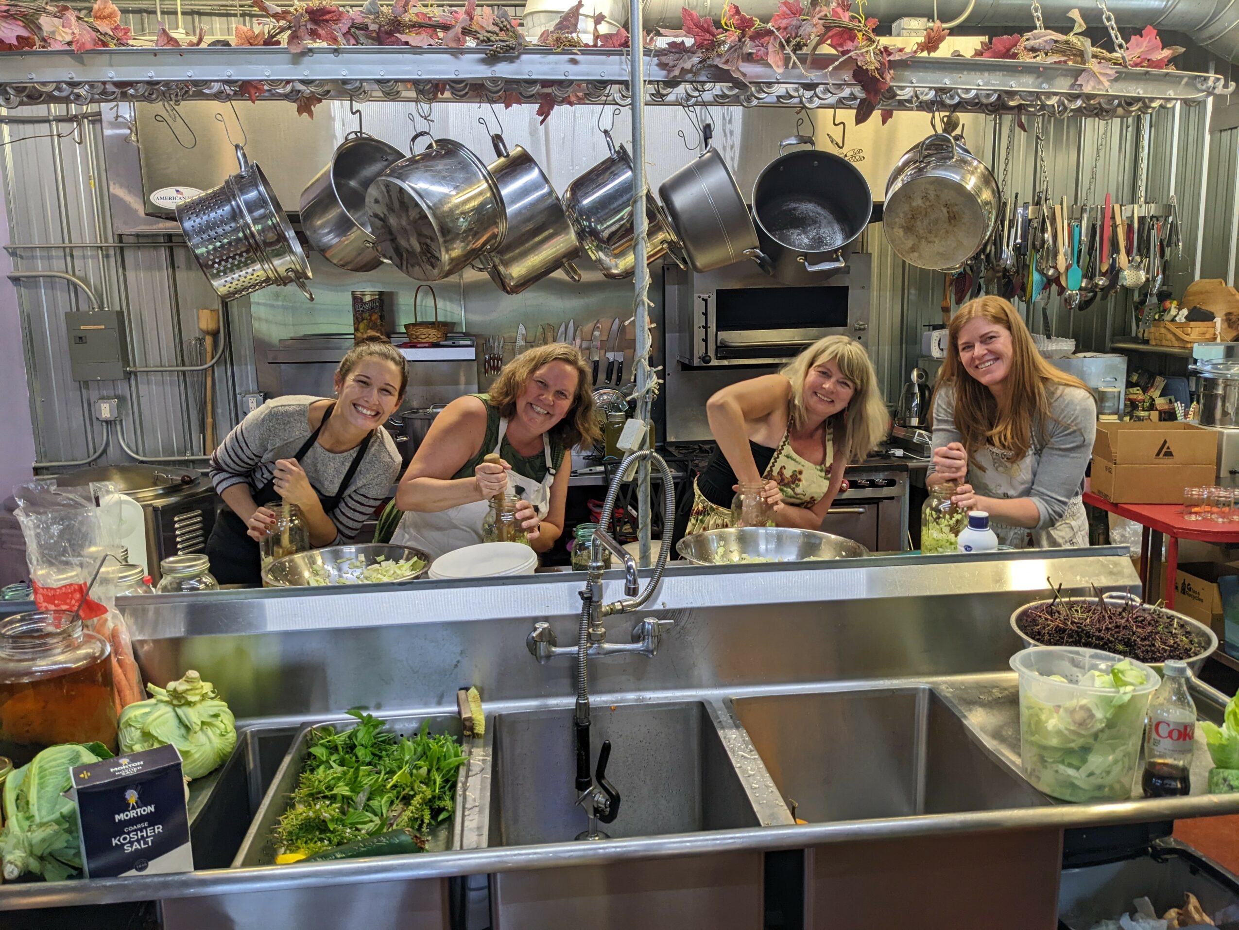 Four women stand behind a large kitchen sink, preparing food with hanging pots and kitchen tools above them.