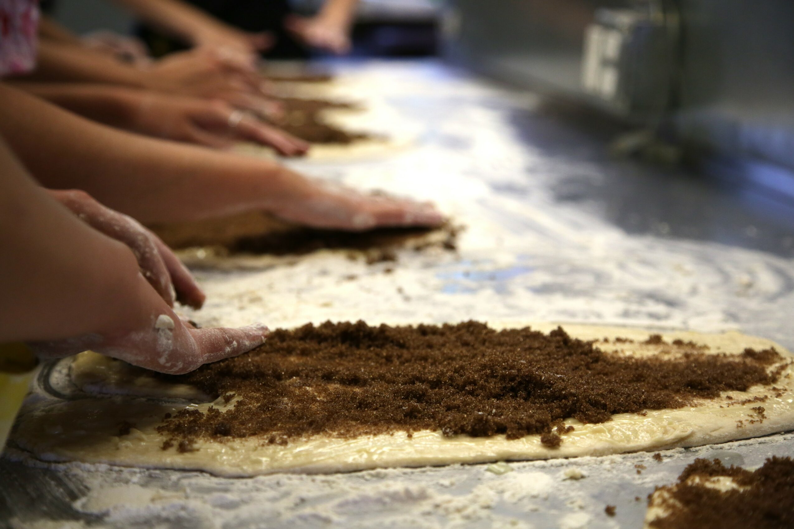 People's hands spreading filling on dough sheet, with more hands in background, in a kitchen or bakery setting.