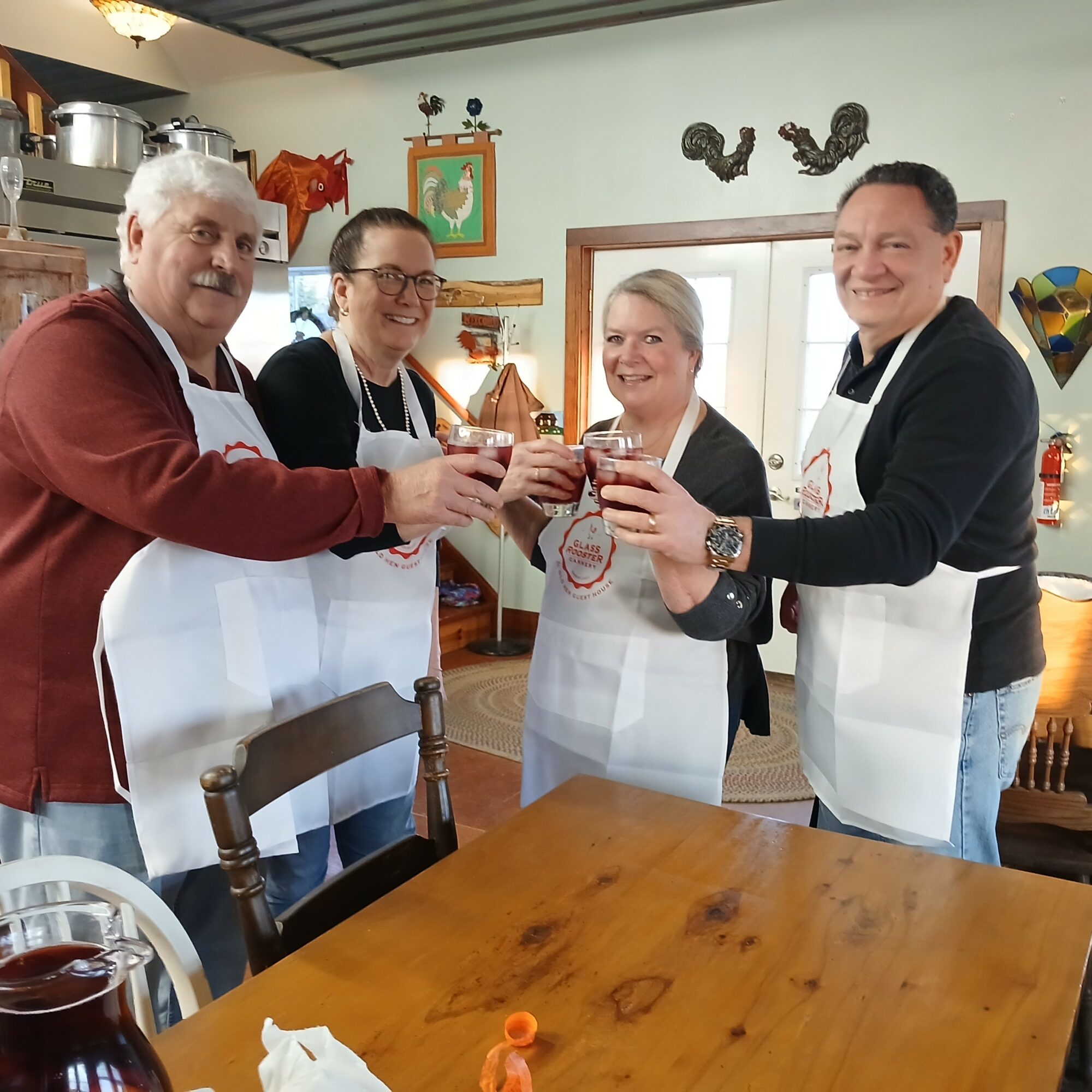 Four people in aprons cheer and toast glasses in a cozy kitchen with decorations and a window behind them.