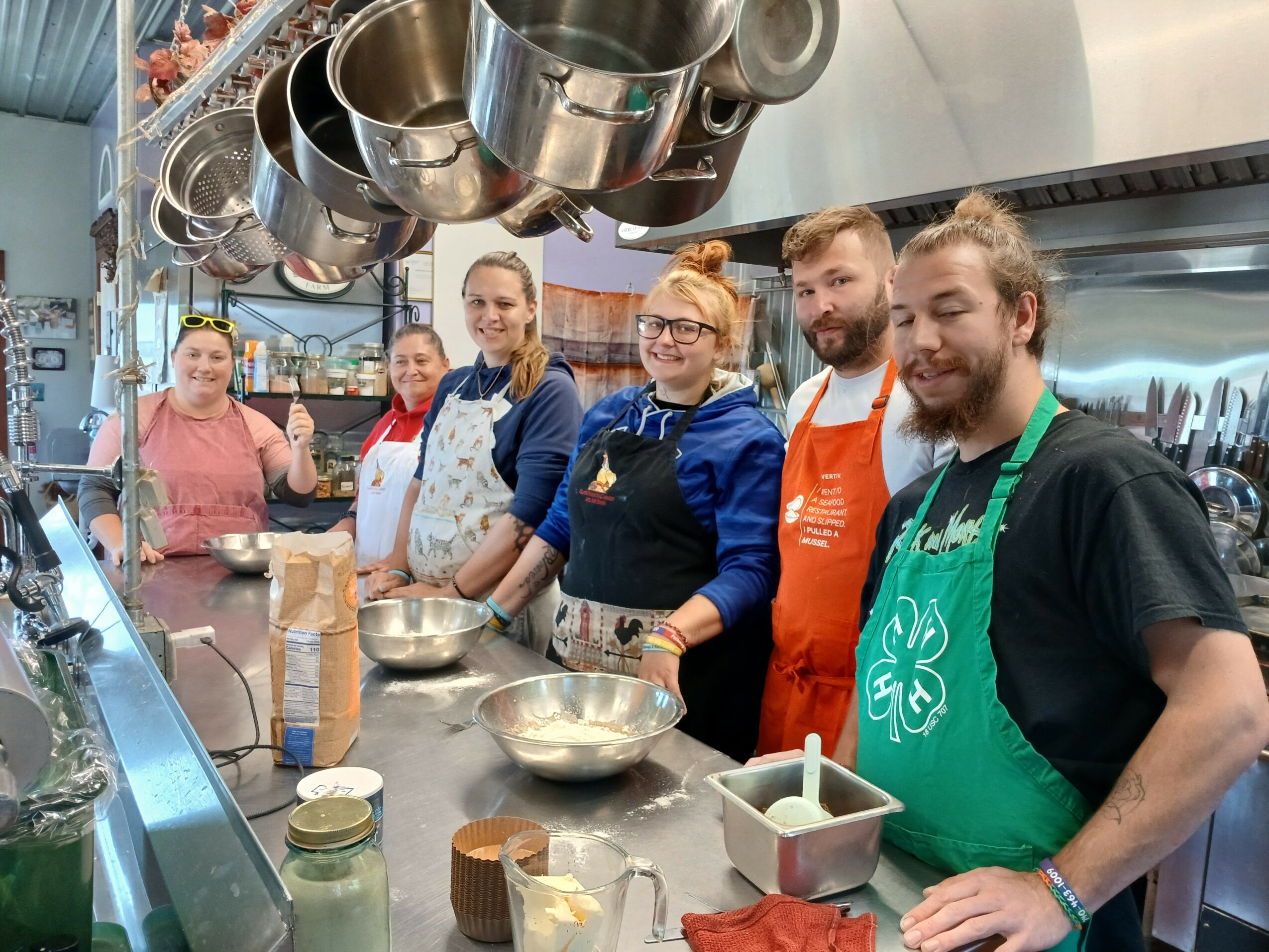Group of six people in a kitchen, wearing aprons, standing behind a counter with bowls and ingredients, smiling.