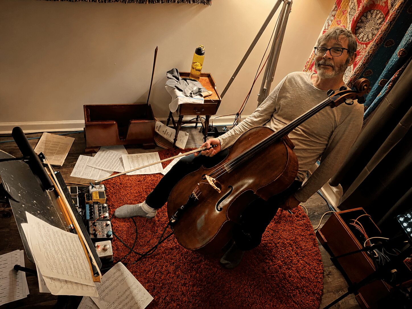 Man sitting on a red rug holding a cello, surrounded by sheet music and musical equipment, in a room with a beige wall.