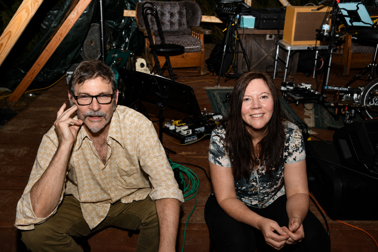 A man with glasses and a woman sit in a music studio with equipment and chairs in the background.