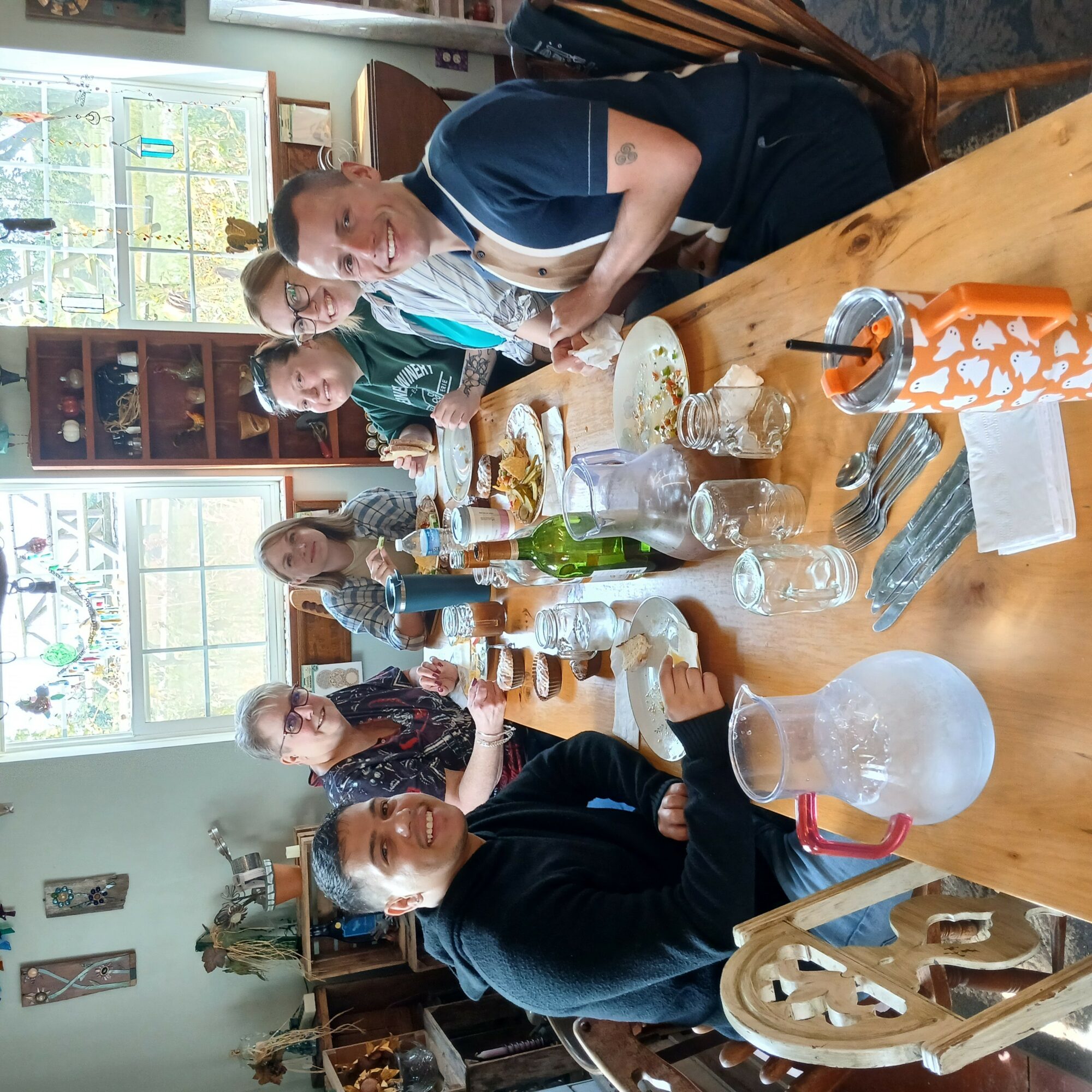 People gathered around a dining table with food and drinks in a room with large windows and wooden furniture.