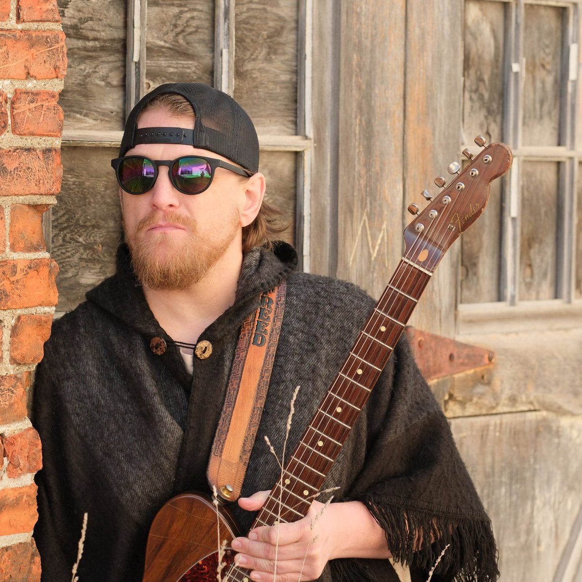 A man with a beard wearing sunglasses, a black cap, and a dark sweater, holding an electric guitar outdoors.