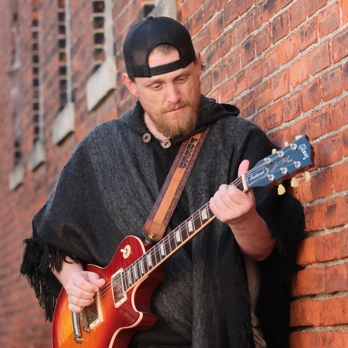Man with beard and cap playing red electric guitar against brick wall, wearing dark poncho.