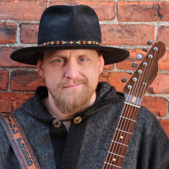 Man with a beard wearing a black hat and dark clothing, holding a guitar, standing in front of a brick wall.